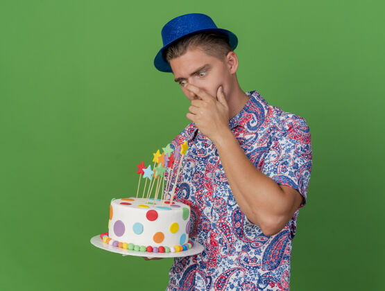 嘴巴惊讶的年轻人戴着蓝帽子拿着蛋糕 看着把手放在嘴上的绿色隔离派对穿上绿色