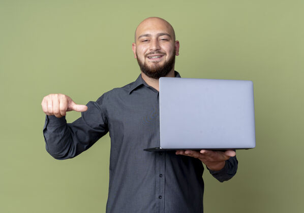姿势微笑的年轻秃顶呼叫中心男子手持笔记本电脑 指着橄榄绿墙上孤立的笔记本电脑笔记本电脑衣服中心