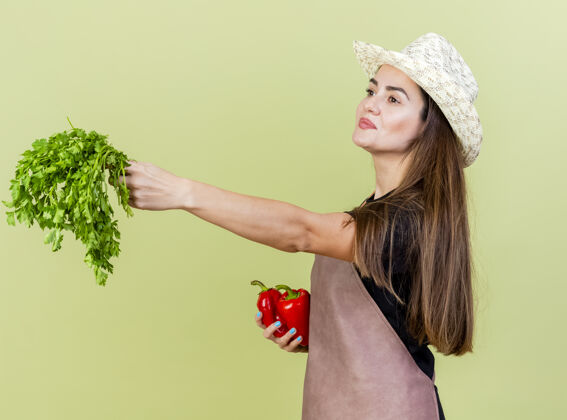 请一个穿着制服的漂亮的园丁女孩戴着园艺帽 手里拿着辣椒 手里拿着香菜 孤零零地站在橄榄绿的一边穿举行制服