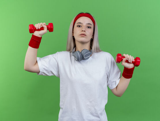 女人自信的年轻运动女性戴着背带 戴着头带 戴着戴着耳机的腕带 举着哑铃看着绿色墙壁上孤立的前方人头带人