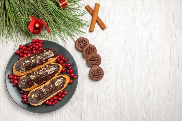 胡椒俯瞰图：灰色盘子上的巧克力和葡萄干饼干交叉着肉桂和松树叶子 白色木桌上放着圣诞玩具肉桂叶子曲奇