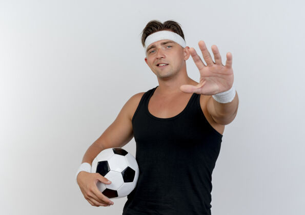 运动高兴的年轻英俊的运动型男子戴着头带和腕带拿着足球 向隔离在白色墙上的前面伸出手来感觉朝向腕带