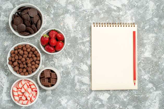 左顶视图碗与糖果草莓巧克力麦片和可可在左边和一个笔记本与红铅笔在右边的灰白色的桌子右纸壁板