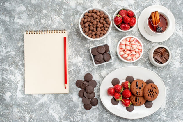 盘子顶视图饼干草莓和圆形巧克力放在椭圆形盘子里碗里糖果草莓巧克力麦片一杯茶和笔记本铅笔放在灰白色的桌子上水果草莓食物
