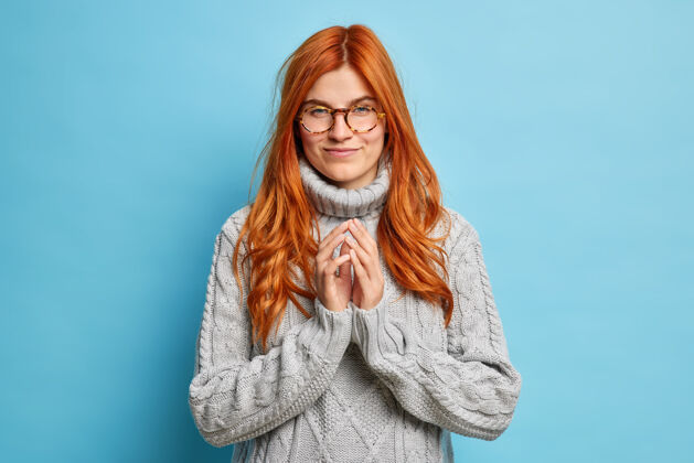 姿势严肃的红发欧洲年轻女子设计的东西尖尖的手指和看起来有意做戴眼镜灰色针织毛衣意图可爱毛衣