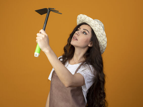 印象年轻的女园丁穿着制服 戴着园艺帽 拿着锄头耙看着橘黄色的墙壁上孤立的复制空间帽子花园锄头