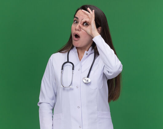 手指兴奋的年轻女医生穿着医用长袍 手持听诊器 用手指对着绿色的相机看年轻女听诊器