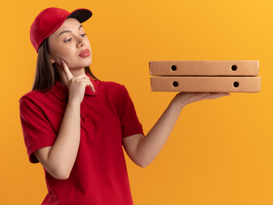 拿着穿着制服的自信漂亮的女送货员把手指放在脸上拿着 看着橙色的比萨饼盒橙色手指脸