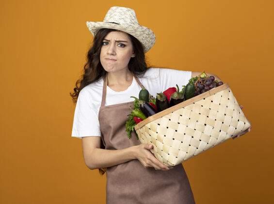 园艺愤怒的年轻女园丁穿着制服 戴着园艺帽 把菜篮子孤立地放在橙色的墙上 留着复制空间制服帽子年轻