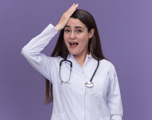 手年轻的女医生穿着带听诊器的医用长袍 手放在头上 看着紫色的相机医学年轻女性