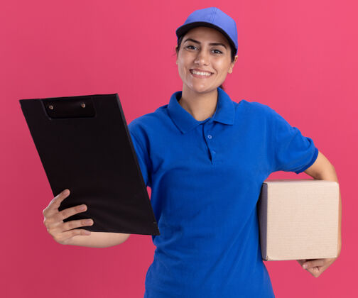 站着面带微笑的年轻送货员 穿着制服 拿着帽子盒 把剪贴板放在粉色的墙上剪贴板拿着姿势