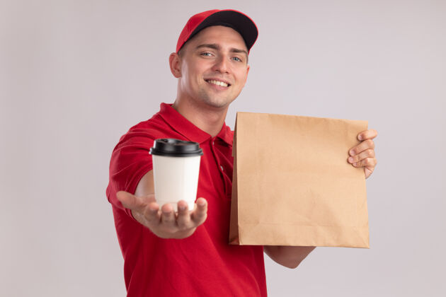 包装面带微笑的年轻送货员穿着制服 戴着帽子 手里拿着纸食品包 手里拿着一杯咖啡 孤零零地站在白色的墙上人咖啡食物