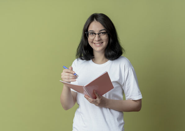 年轻微笑着的年轻漂亮的高加索女孩戴着眼镜拿着笔和笔记本孤立在橄榄绿的背景与复印空间眼镜微笑笔记