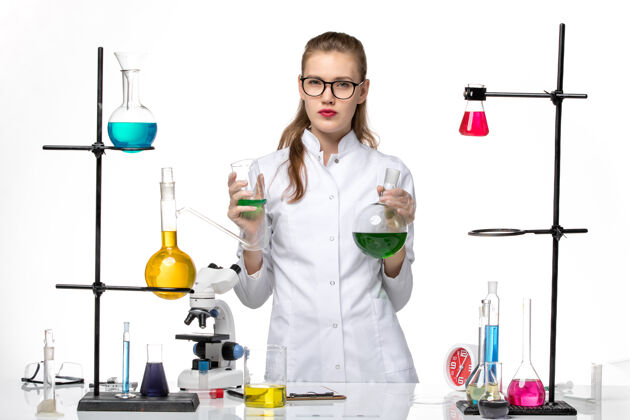 医学前视图穿着医疗服的女化学家拿着浅白色背景上带绿色溶液的烧瓶化学大流行性冠状病毒烧瓶化学人