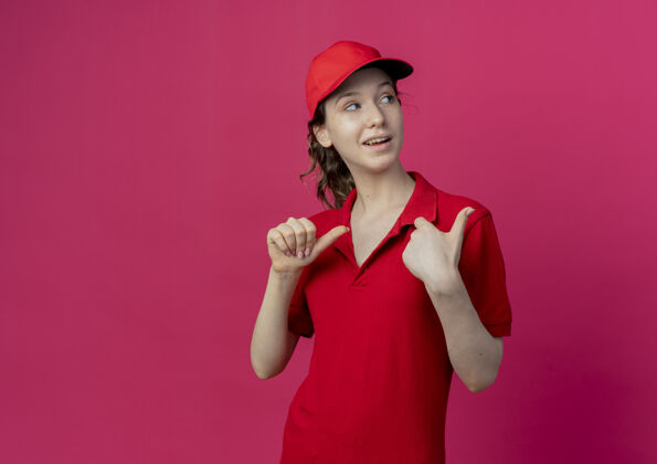 深红色令人印象深刻的年轻漂亮的送货女孩穿着红色制服 戴着帽子 看着和指着一边孤立的深红色背景与复制空间制服壁板印象