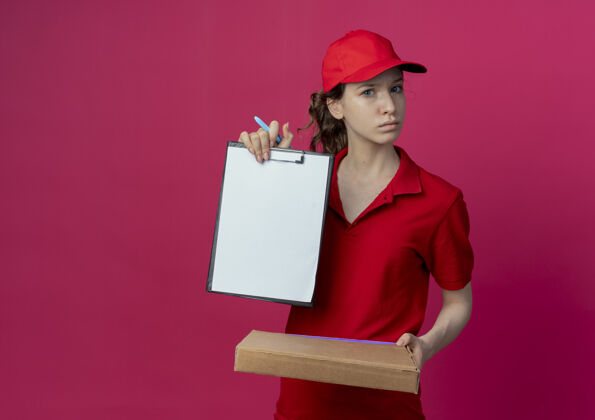 漂亮令人印象深刻的年轻漂亮的送货女孩在红色制服和帽子举行比萨饼包装笔和显示剪贴板上的深红色背景与复制空间隔离背景披萨深红色