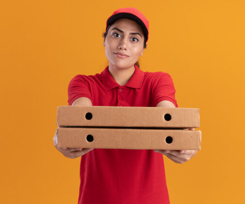 拿着身着制服 头戴鸭舌帽 手持披萨盒的年轻送货员高兴地站在橙色的墙上递送年轻人穿着