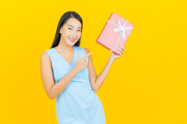 人肖像美丽的亚洲年轻女子微笑与黄色墙上的红色礼盒女士时尚年轻