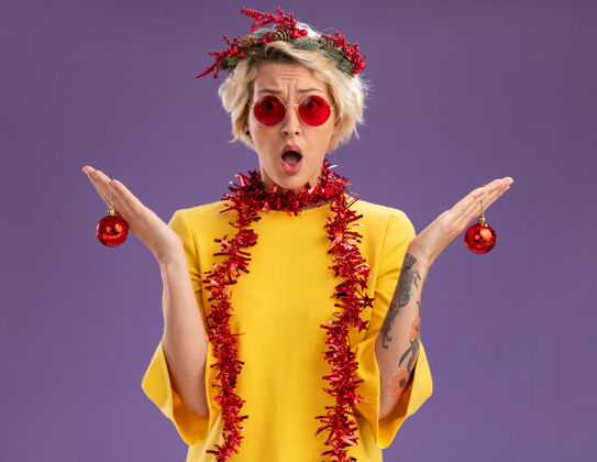 花环笨手笨脚的金发女郎戴着圣诞花环 脖子上戴着金属丝花环 手里拿着圣诞饰品 看着紫色背景上孤立的摄像机周围不知所措抱着