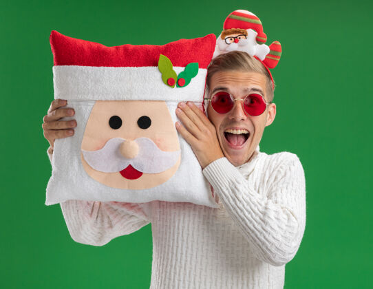 抱给人印象深刻的年轻帅哥戴着圣诞老人的头带戴着眼镜抱着圣诞老人的枕头靠近头部看着隔离在绿色背景上的相机圣诞快乐头绿色
