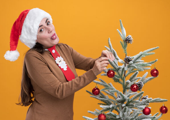 立场快乐的年轻漂亮女孩戴着圣诞帽打着领带站在圣诞树旁 舌头孤立在橙色的背景上帽子圣诞树舌头