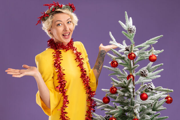 圣诞快乐年轻的金发女郎戴着圣诞花环 脖子上戴着金箔花环 站在装饰好的圣诞树旁 看着镜头 紫色背景上空空的双手与世隔绝花圈年轻金发