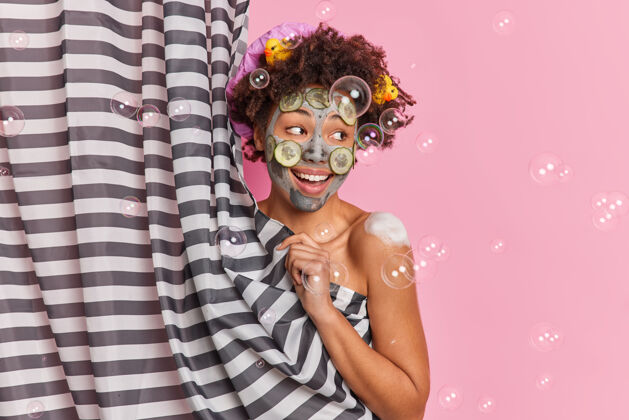 健康快乐的卷发女人用黄瓜片涂上粘土面膜享受淋浴正面摆出半裸的姿势窗帘后有橡皮鸭在头发上隔离粉色背景旁边赤膊上阵粉色