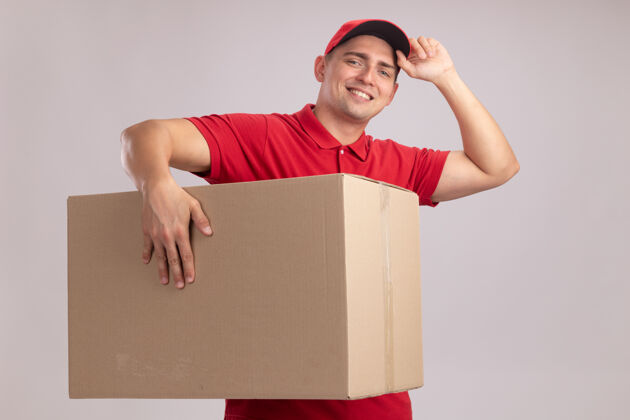 穿微笑的年轻送货员穿着制服 戴着帽子拿着大箱子 帽子孤立在白色的墙上微笑拿着年轻