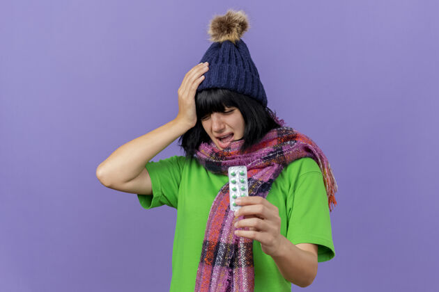 年轻人带着冬天帽子和围巾的疼痛的年轻病妇手放在头上拿着一包胶囊闭着眼睛头痛 隔离在紫色的墙上 留着复印空间站头痛保持