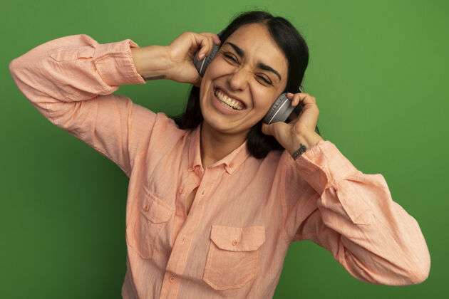 女孩带着微笑的年轻漂亮女孩穿着粉色t恤 头戴耳机隔离在绿色的墙上戴上微笑年轻