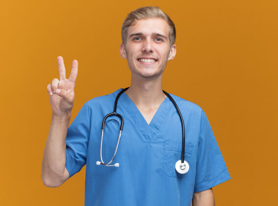 年轻微笑着的年轻男医生穿着医生制服 带着听诊器 在橙色的墙上显示出和平的姿态佩戴微笑和平