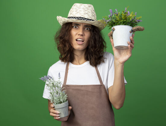 花迷茫的年轻女园丁穿着制服戴着园艺帽捧着鲜花在花盆里隔离在绿茵场上女性制服园艺