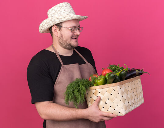 花园高兴的年轻男园丁戴着园艺帽 手拿蔬菜篮子看着隔离在粉红色墙上年轻篮子帽子