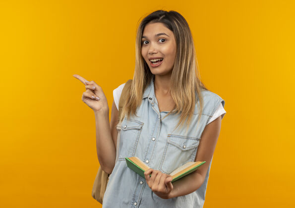 指着快乐的年轻漂亮的女学生 背着书包 拿着打开的书 指着橘子树上孤立的一面袋子快乐橙色