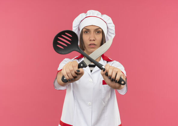 不愉快穿着厨师制服的年轻女厨师拿着抹刀和小刀 对着隔离在粉色背景上有复印空间的摄像机做着“不”的手势厨师抹刀制服