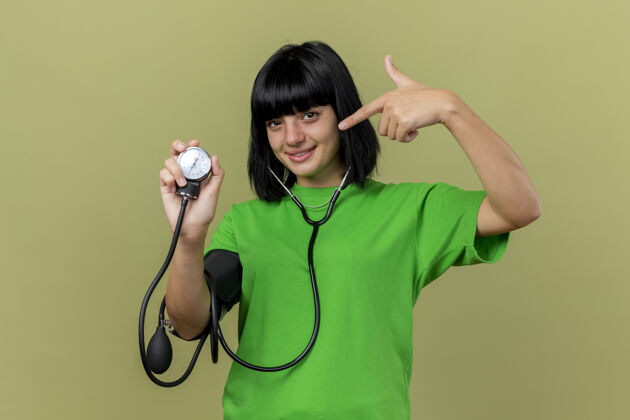 疾病戴着听诊器的年轻病妇高兴地看着血压计指着它 在橄榄绿的墙壁上看前面的隔离复制空间站立市民人