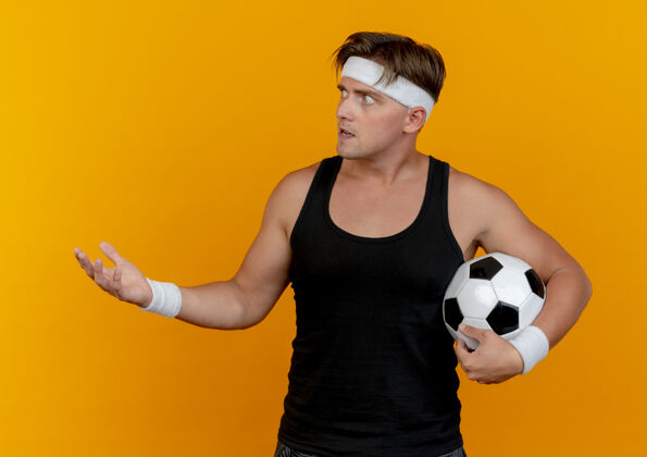 运动令人印象深刻的年轻英俊的运动男子戴着头带和腕带拿着足球看在一边 并显示空手孤立在橙色空持有穿