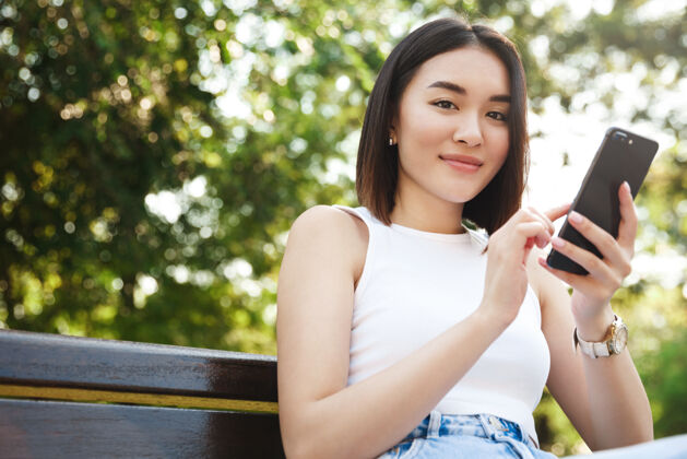 亚洲时尚的亚洲女孩坐在公园里用智能手机对着镜头微笑微笑聊天女孩