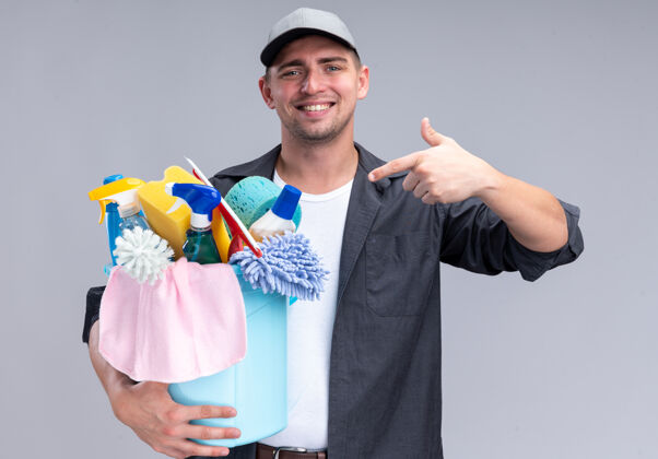 点微笑着的年轻帅气的清洁工穿着t恤 戴着帽子 指着隔离在白墙上的一桶清洁工具T恤家伙微笑