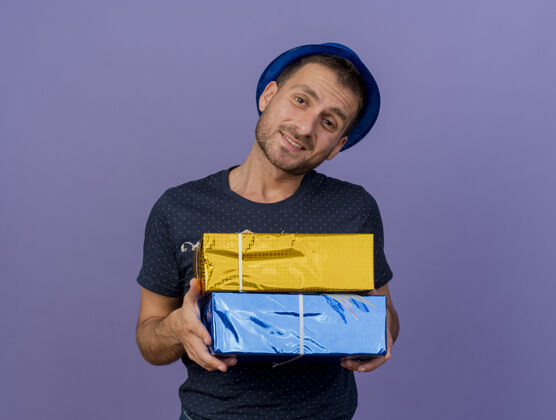 蓝色戴着蓝色帽子的白种帅哥拿着礼盒 看着相机在紫色背景上的复制空间盒子副本紫色