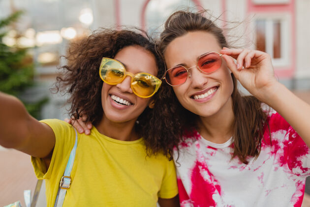 太阳镜五颜六色的照片 快乐的年轻女孩朋友们微笑着坐在街上用手机自拍 女人们在一起玩得很开心女人欢呼非洲裔