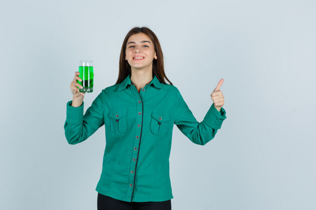 时尚年轻女孩穿着绿色的上衣 黑色的裤子 手里拿着一杯绿色的液体 竖起大拇指 看上去很开朗 眼前一亮青年女士玻璃