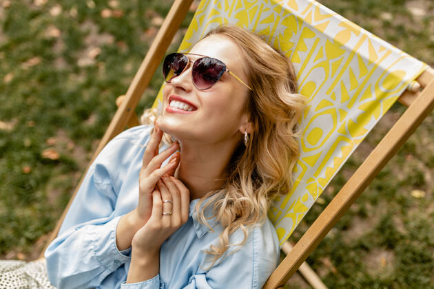 穿迷人的金发微笑的女人坐在躺椅上穿着夏天的衣服快乐太阳镜放松