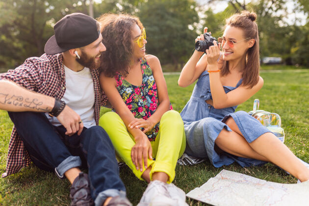 相机快乐的年轻朋友们一起坐在公园里 男女同乐 缤纷的夏日潮人时尚风格 带着相机旅行 笑口常开黑色青春人