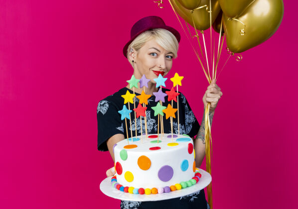 伸展高兴的年轻金发派对女孩戴着派对帽看着相机拿着气球和伸展生日蛋糕与星星对着相机在深红色背景与复制空间隔离帽子金发派对