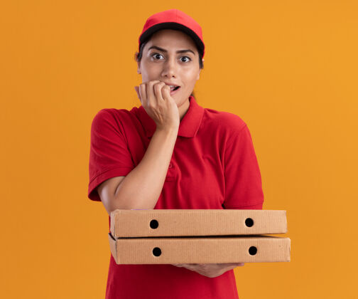 女孩令人印象深刻的年轻送货女孩穿着制服和帽子举行比萨饼盒咬钉子隔离在橙色墙上披萨盒子年轻
