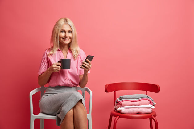 年龄高兴的五十岁的老太太拿着手机和茶杯在社交网络上冲浪 在家里闲暇时独自坐在舒适的椅子上享受在线交流生活方式的概念成熟电话茶