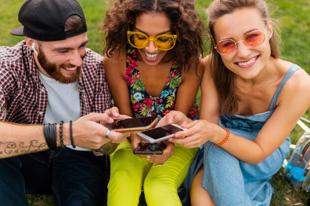 多彩快乐的年轻朋友们坐在公园里用智能手机微笑 男人和女人一起玩朋友女人微笑
