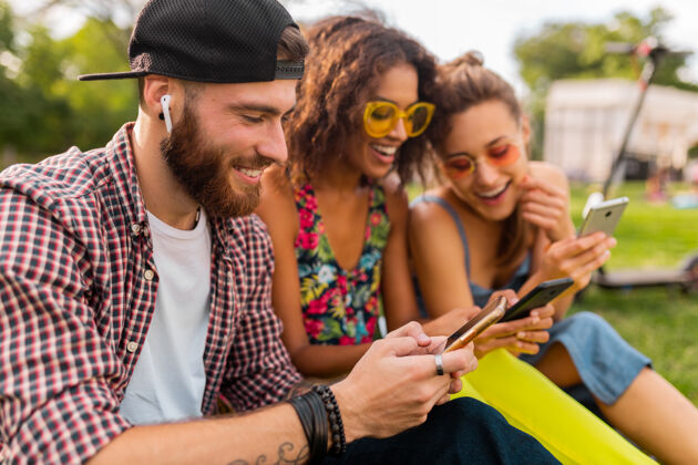 眼镜快乐的年轻朋友们坐在公园里用智能手机微笑 男人和女人一起玩兴奋多样性女人