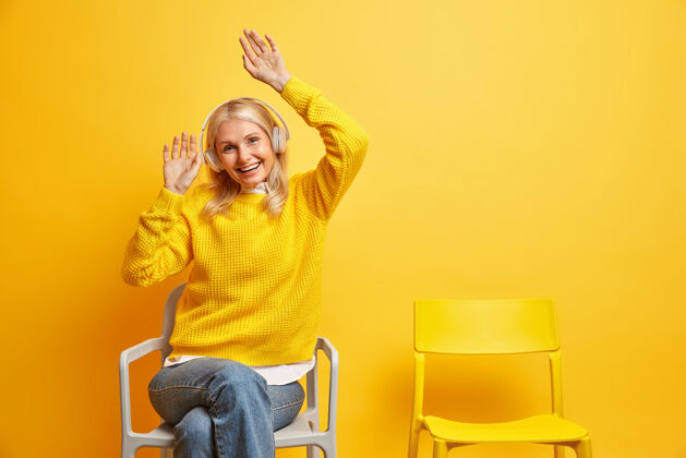 享受人们休闲娱乐的概念有趣的金发老妇人举起双臂坐在舒适的椅子上 通过无线耳机收听音频轨道成人音频耳机